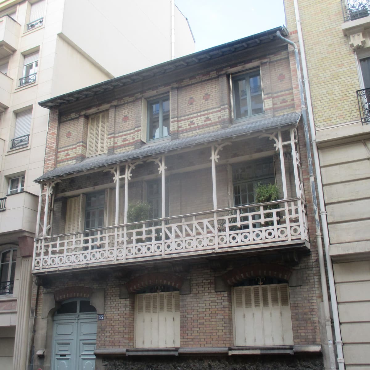 Maison étroite de briques claires à bandes colorées, avec un balcon couvert en bois découpé au premier étage, entre deux immeubles.